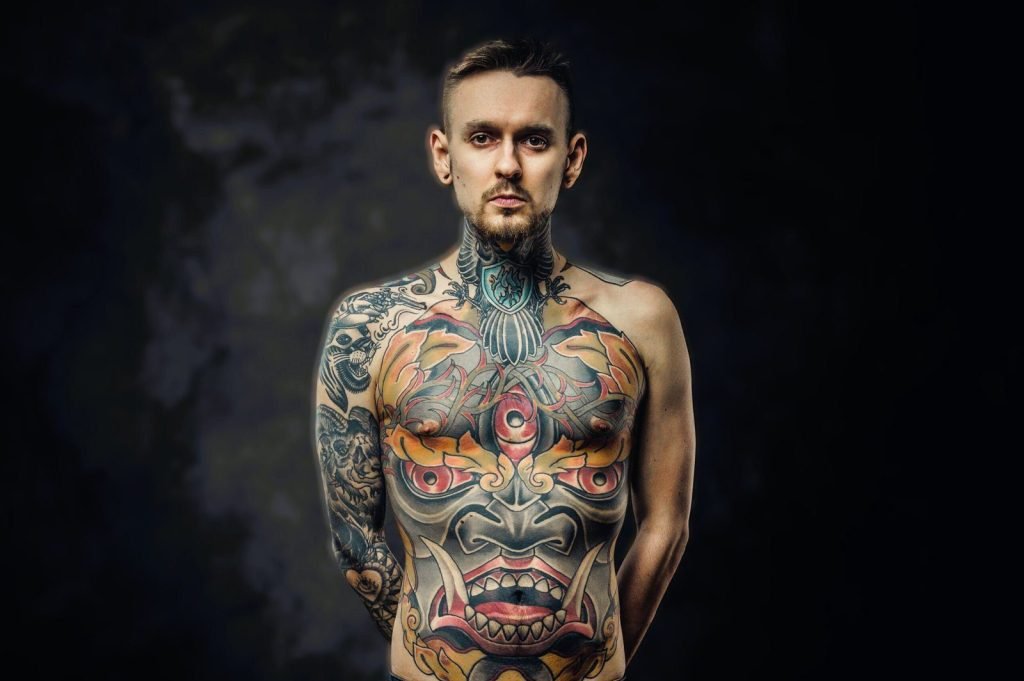 Assertive tattoo artist posing in a dark studio with a half-naked body wearing jeans, tattooed in a japanese irezumi style, looking cool and confident.